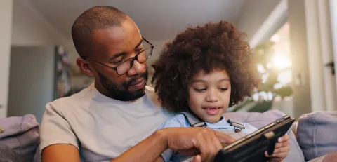 An adult with a child on his knee both engaging with a technological device