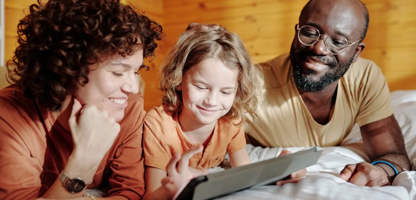 2 adults smiling whilst watching a young person use an electronic device