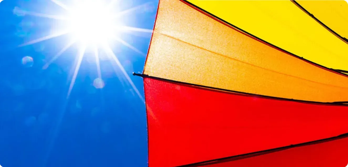 An image of the sun in a blue sky shining down on a red, orange and yellow umbrella